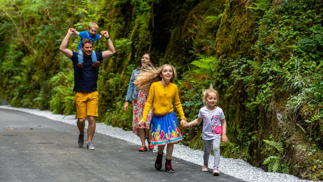 A family of five walks along a paved path through lush greenery, with two girls in front holding hands and parents with a toddler and young boy behind them.