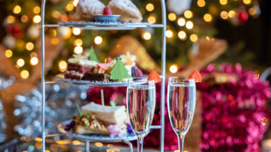 A three-tiered tray with sandwiches, pastries, and desserts sits beside two glasses of sparkling wine, with festive holiday lights in the background.