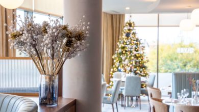 A glass vase with decorative white berries is on a counter in a restaurant, with a decorated Christmas tree and set dining tables in the background.