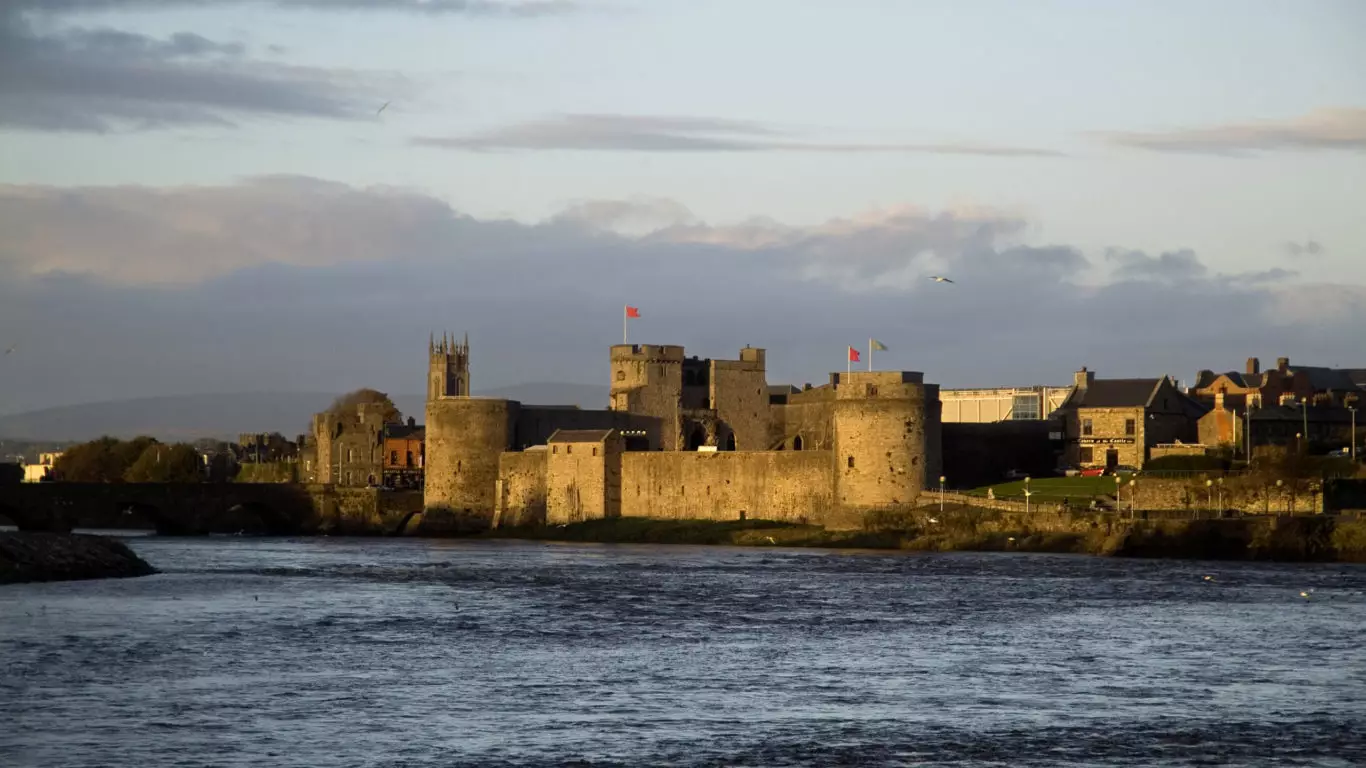 A medieval stone castle with round towers and battlements stands beside a river under a partly cloudy sky at sunset.