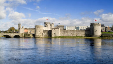 Stone castle with round towers and flags, located beside a river on a sunny day with blue sky and scattered clouds; bridge to the left and buildings in the background.