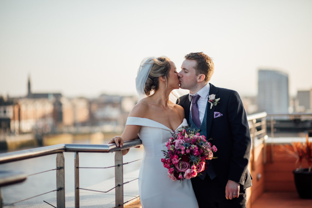 Bride and groom kiss on an outdoor balcony, dressed in wedding attire; the bride holds a bouquet of pink flowers. City buildings are visible in the blurred background.
