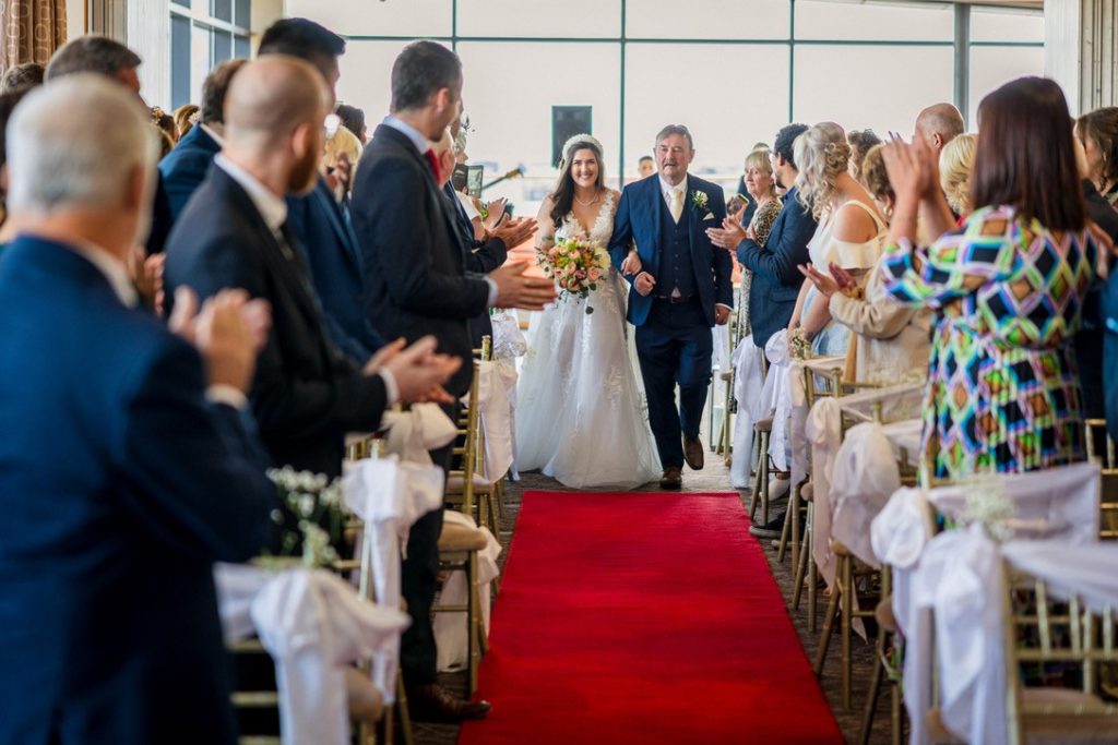 A bride in a white dress walks down a red carpeted aisle with an older man, surrounded by seated and standing guests applauding.