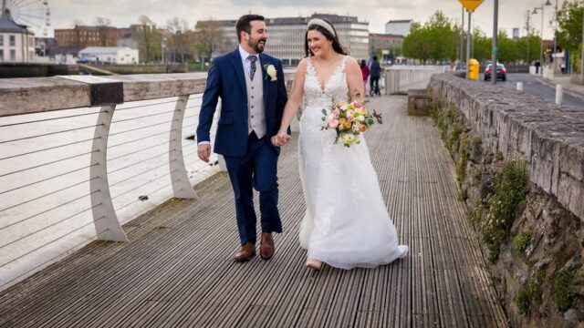 A bride and groom walk hand in hand on a riverside boardwalk, both smiling. The bride holds a bouquet of flowers and wears a white dress; the groom wears a blue suit.
