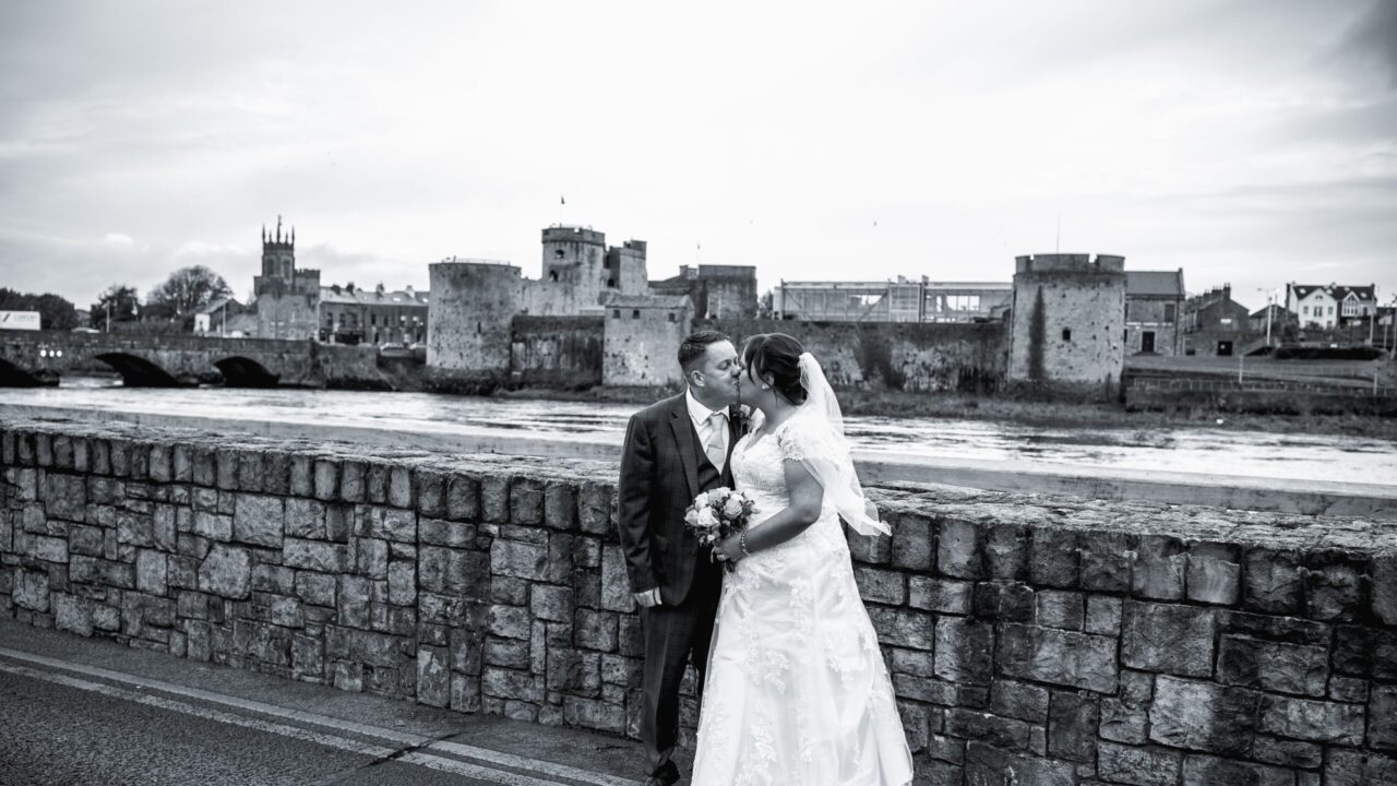 Bride and groom kiss while standing by a stone wall, with a historic castle and river in the background.