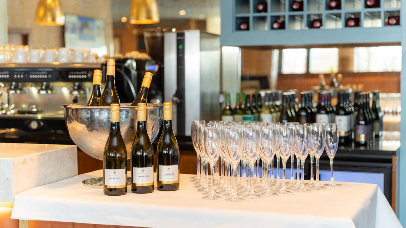 A table with bottles of white wine in a metal bucket, several empty wine glasses, and more wine bottles in the background at a bar or restaurant.