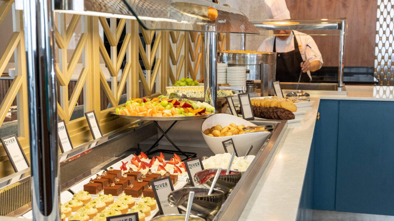 Buffet counter with assorted desserts, fruit platter, and other food items; a chef in uniform stands behind the counter.