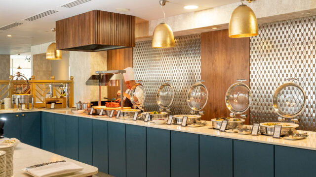 A chef stands behind a buffet counter with various covered dishes in a modern, well-lit restaurant setting.