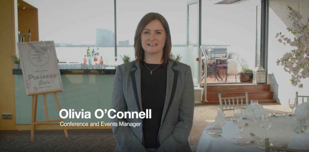 A woman in business attire stands in a bright event space near a set dining table and a bar sign, with “Olivia O’Connell, Conference and Events Manager” overlaid on the image.