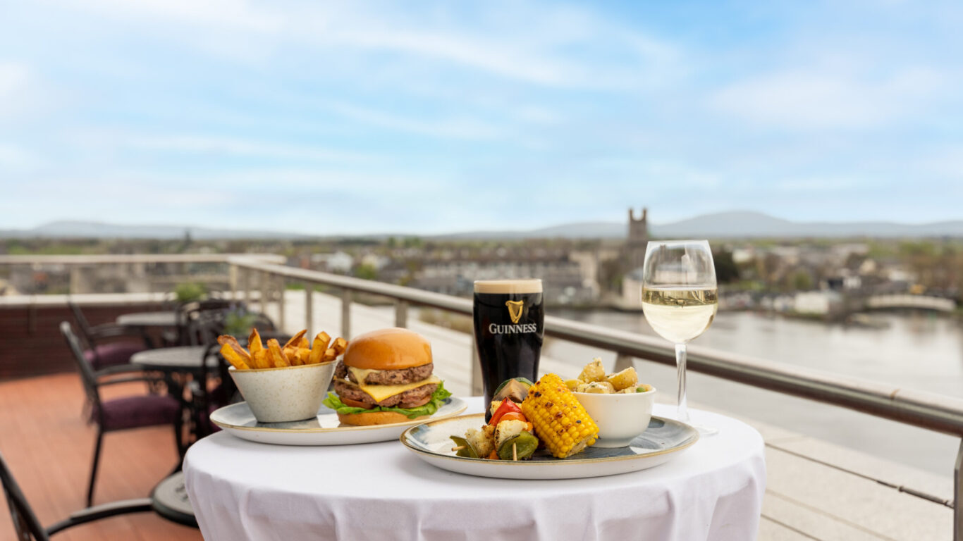 A table on a balcony overlooking a river, set with fries, a burger, a Guinness pint, a glass of white wine, corn on the cob, and a bowl of mixed vegetables.