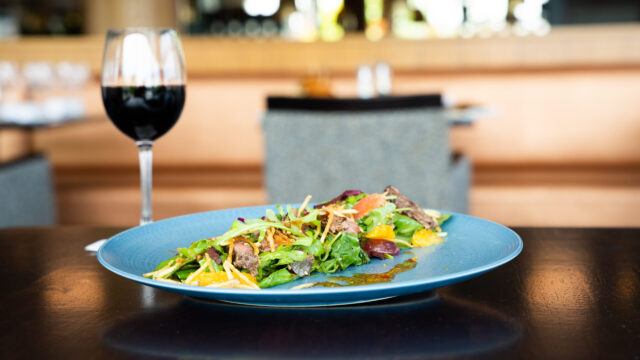 A plate of salad with sliced steak and vegetables on a blue dish, next to a glass of red wine on a dark table in a restaurant setting.