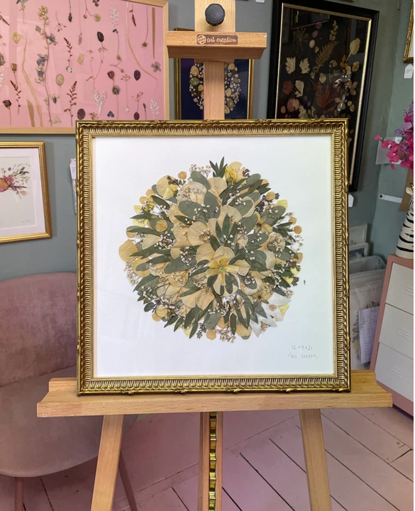 Framed botanical artwork featuring a circular arrangement of dried leaves and flowers displayed on an easel in an art studio.