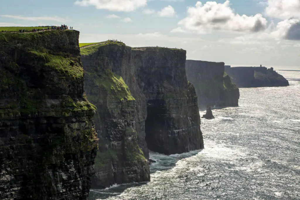 Cliffs of Moher rise steeply above the Atlantic Ocean under a partly cloudy sky, with people on the grassy edge—an inspiring view perfect for those seeking memorable Autumn Hotel Breaks nearby.