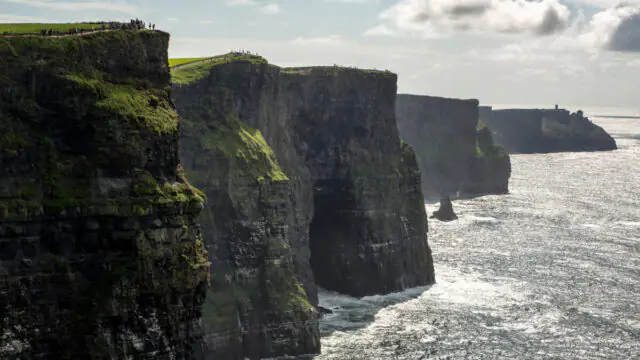 Cliffs of Moher rise steeply above the Atlantic Ocean under a partly cloudy sky, with people on the grassy edge&mdash;an inspiring view perfect for those seeking memorable Autumn Hotel Breaks nearby.