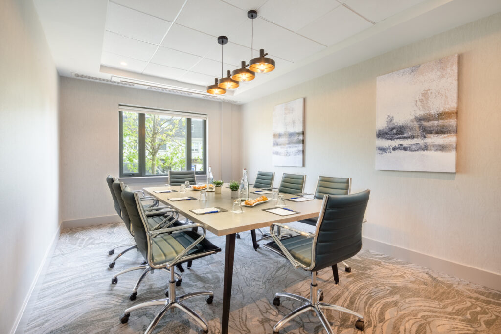 A modern conference room with a long table, eight chairs, notepads, water bottles, snacks, abstract wall art, and a window letting in natural light.