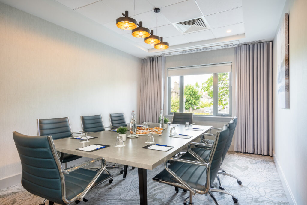 A modern conference room with eight chairs around a rectangular table set with notepads, pens, water bottles, snacks, and a window with a view of greenery.