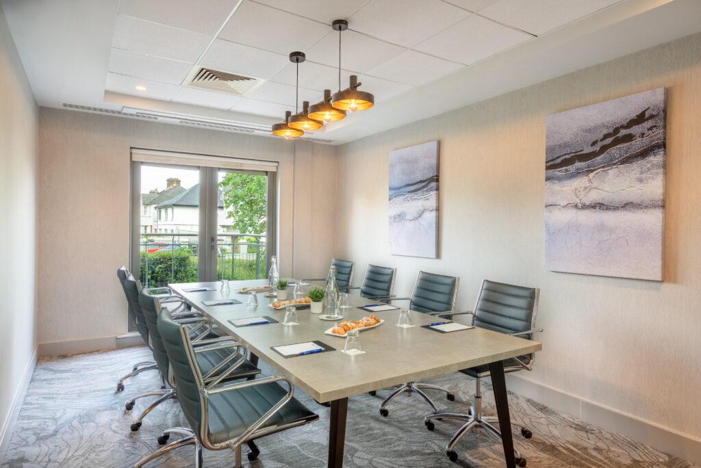 A modern conference room with a long table, eight chairs, snacks, bottled water, notepads, and pens, with large windows and abstract art on the walls.