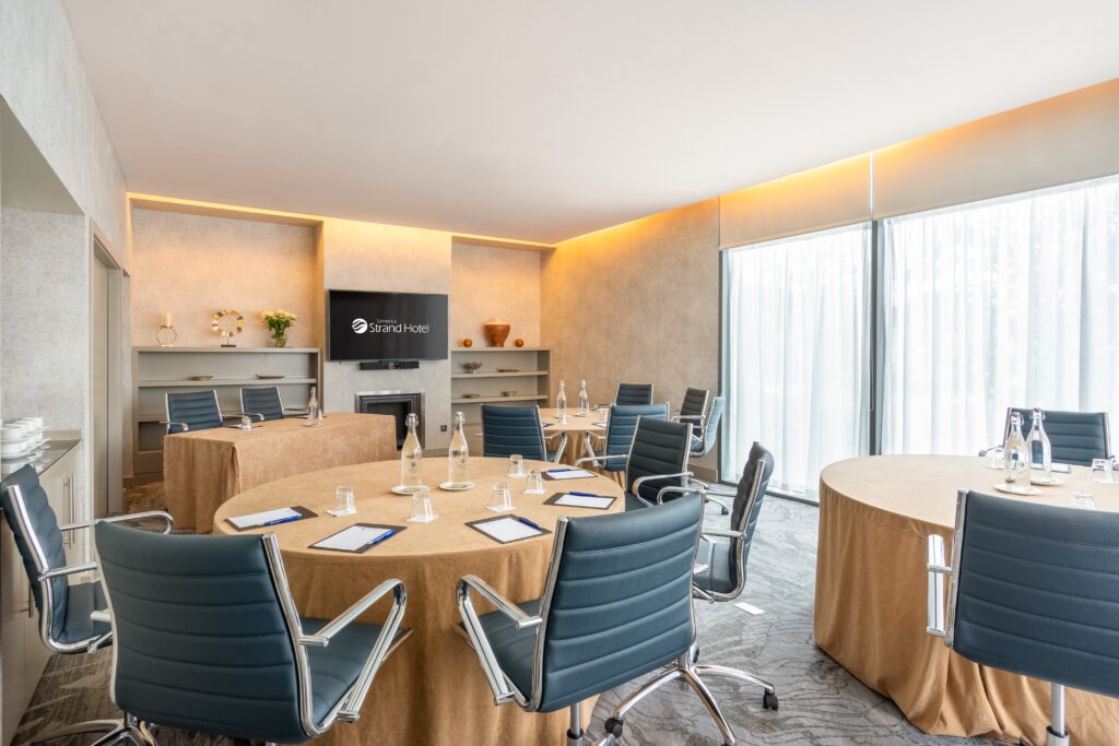 Modern conference room with round tables, black chairs, notepads, and water bottles. Large windows with sheer curtains provide natural light. A screen displays 