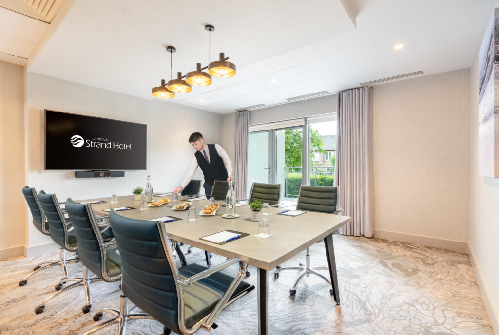 A hotel staff member sets up refreshments on a conference table in a modern meeting room with eight chairs, a large window, and a wall-mounted TV displaying 
