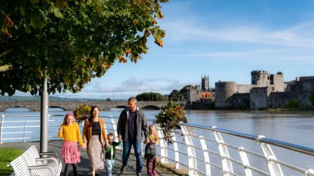 A family of five strolls along a riverside promenade with a castle and stone bridge across the water under sunny skies, enjoying the perfect setting for memorable autumn hotel breaks.