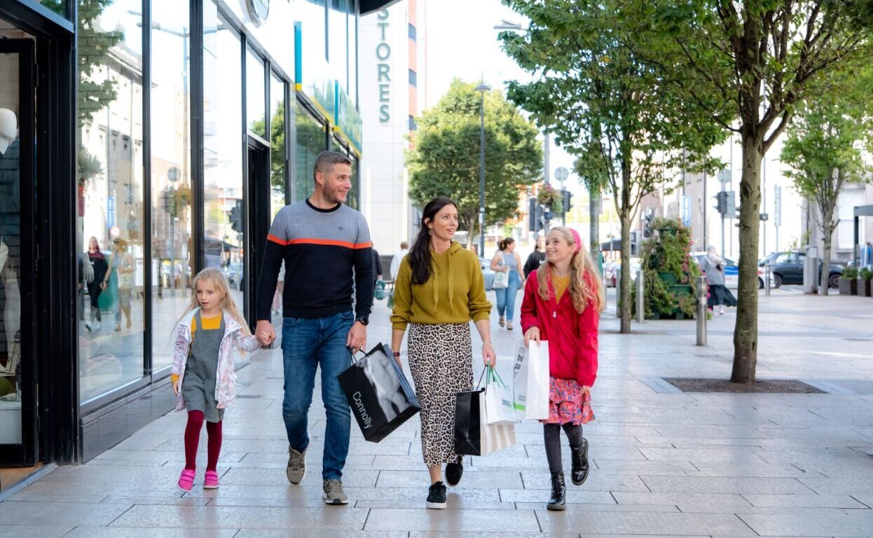 A family of four walks along a shopping street, carrying shopping bags and dressed in casual clothing, enjoying the crisp air and vibrant scenery—perfect for those seeking inspiration for autumn hotel breaks.