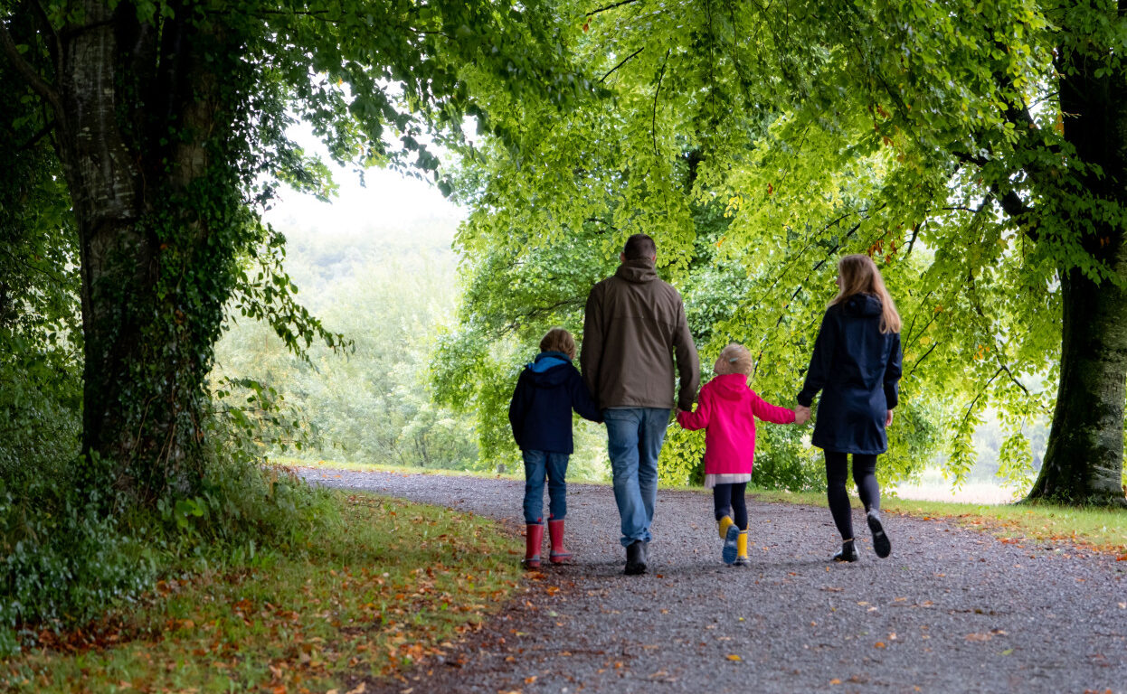 Two adults and two children walk on a gravel path through a green, leafy park, holding hands and wearing jackets and boots—enjoying quality time together during their Autumn Hotel Breaks.