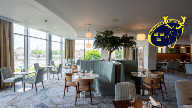 Modern restaurant interior with light blue seating, round tables set for dining, large windows, and the Munster Rugby logo in the upper right corner.