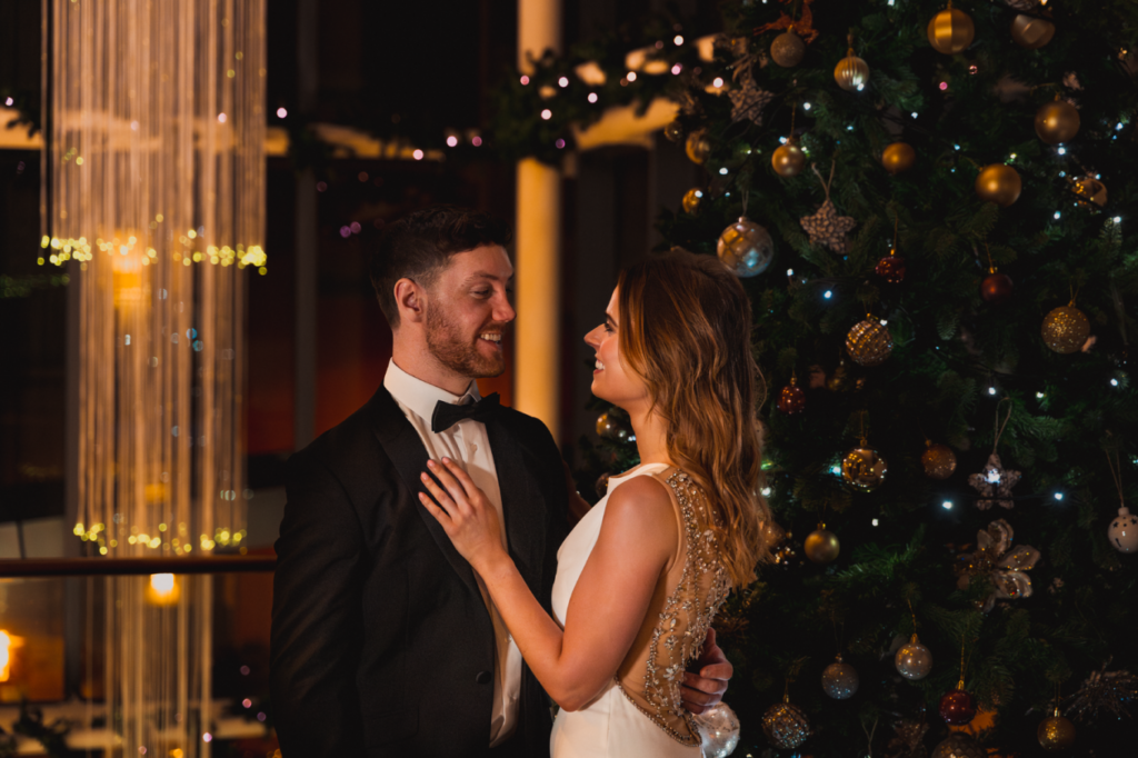 A man in a tuxedo and a woman in a white dress stand together beside a decorated Christmas tree, looking at each other and smiling in a warmly lit room.