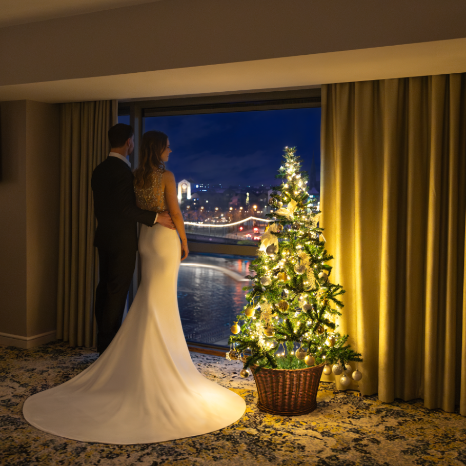 A couple dressed in formal attire stands by a window overlooking a city at night, next to a decorated Christmas tree inside a room.