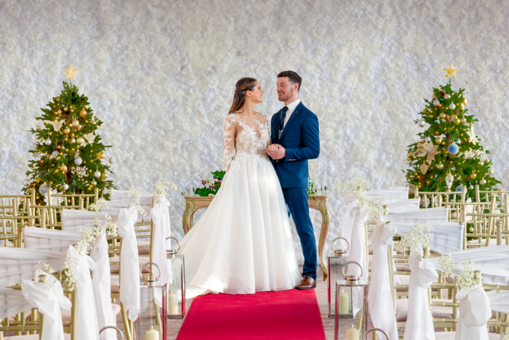 A bride and groom stand together at the front of a decorated wedding aisle with two Christmas trees behind them.