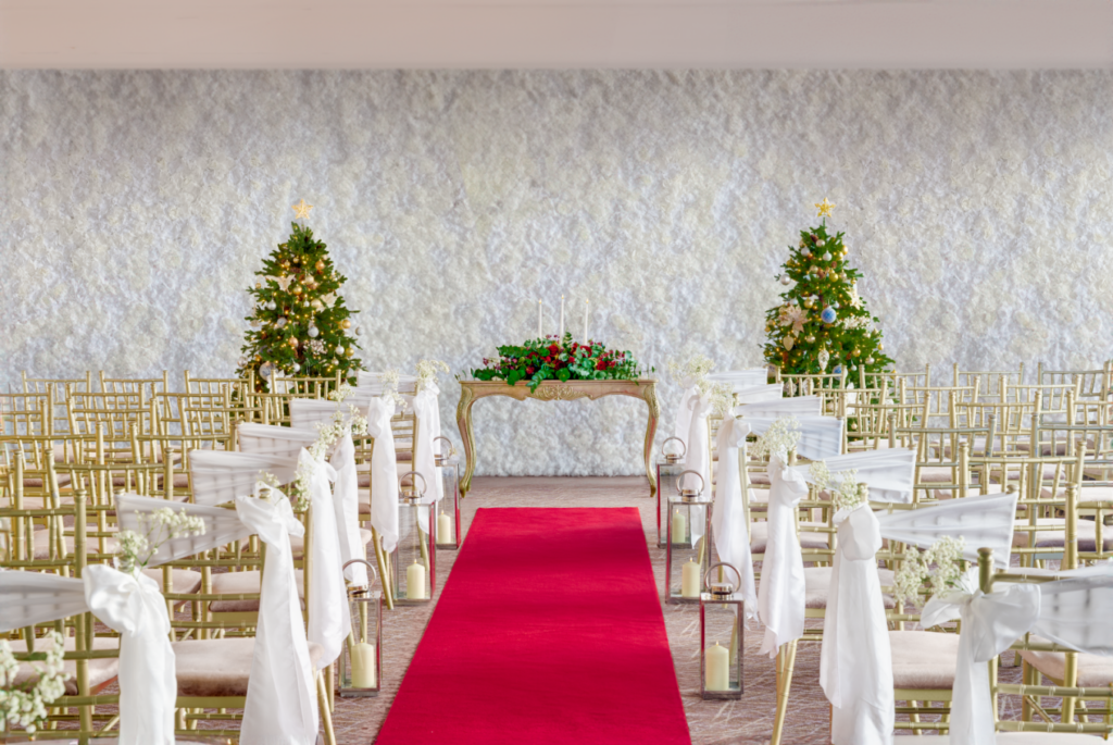 Rows of gold chairs with white fabric line a red aisle runner facing a table with floral decor and two decorated Christmas trees in a bright indoor wedding setting.