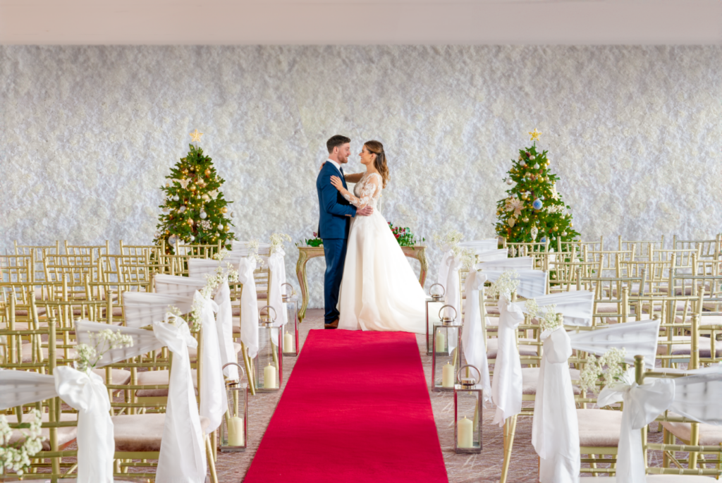 A bride and groom stand together at the end of a red carpet in a decorated venue with rows of chairs and two Christmas trees in the background.