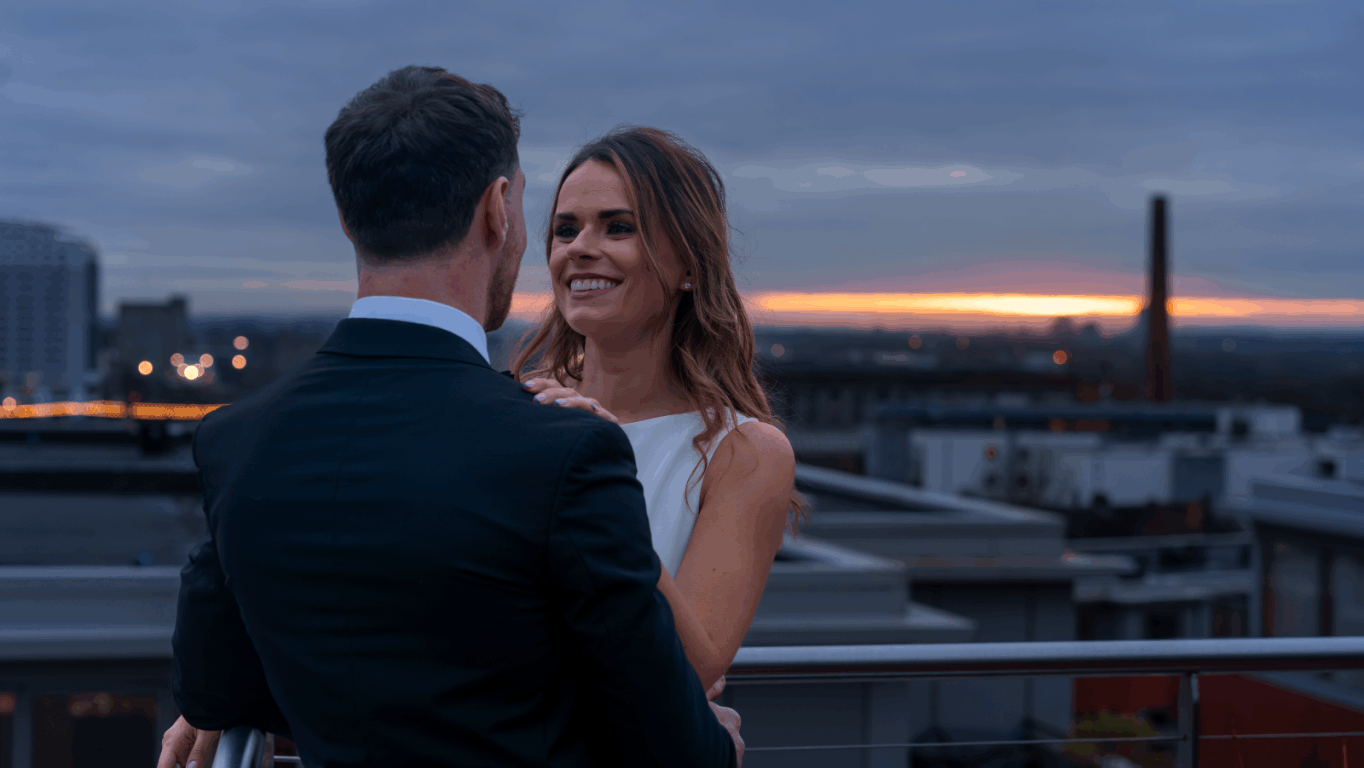 A couple dressed formally stands on a rooftop at sunset, with the cityscape and a cloudy sky in the background.