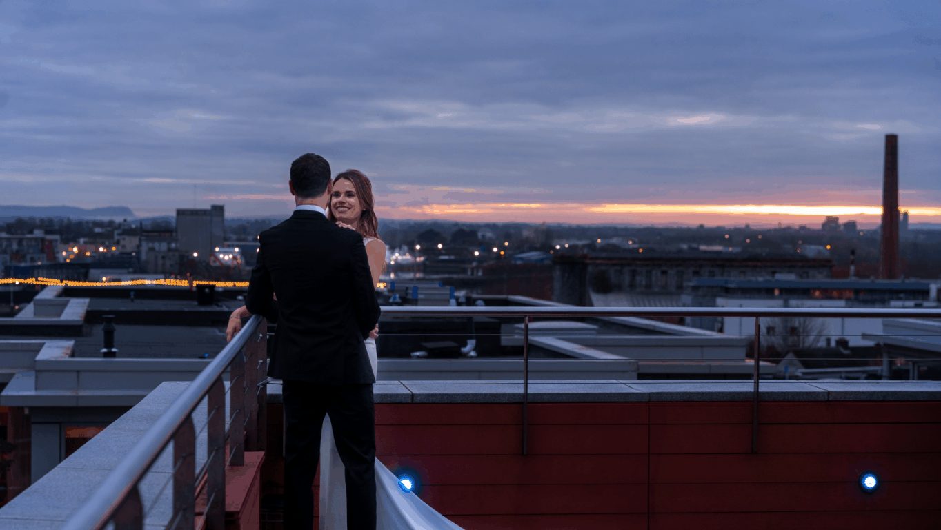 A bride and groom stand together on a rooftop deck at sunset, overlooking a cityscape with buildings and a cloudy sky in the background.
