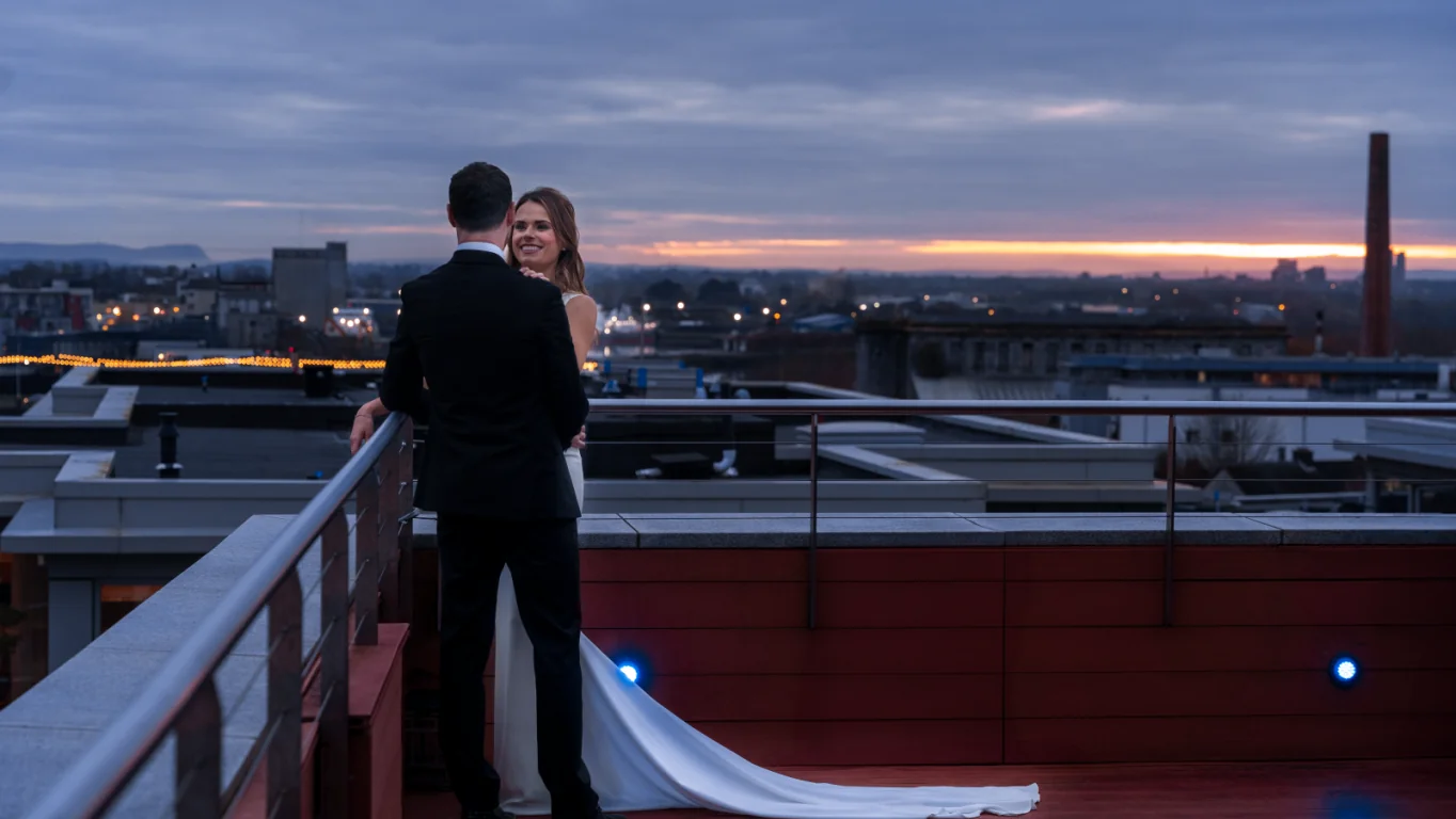 A bride and groom stand together on a rooftop deck at sunset, overlooking a cityscape with buildings and a cloudy sky in the background.