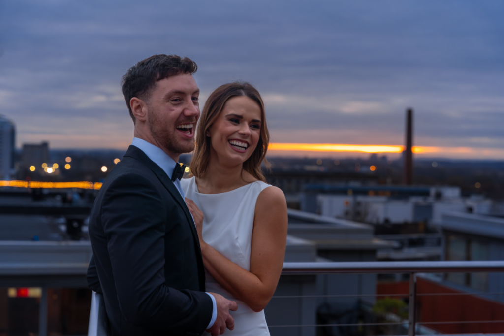 A man in a suit and a woman in a white dress smile together on a rooftop at sunset, with city buildings and lights in the background.