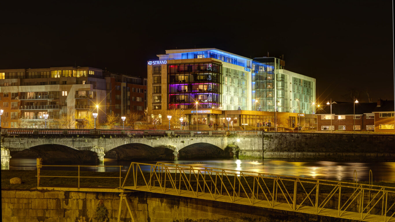 A modern riverside hotel and nearby buildings are illuminated at night, with a stone bridge and water reflecting the lights in the foreground.