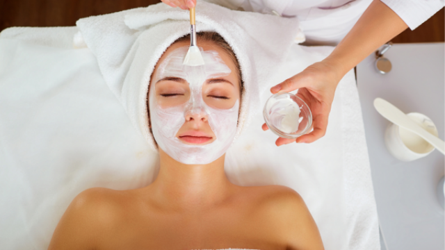 A person lies with eyes closed while a facial mask is applied to their face using a brush by another person in a spa setting.