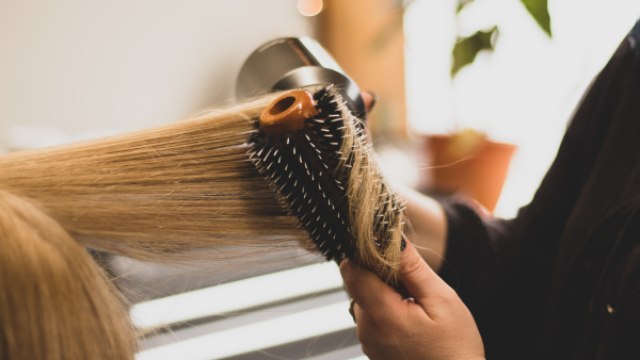 A person uses a round brush and hair dryer to style long, straight blonde hair in a well-lit indoor setting.