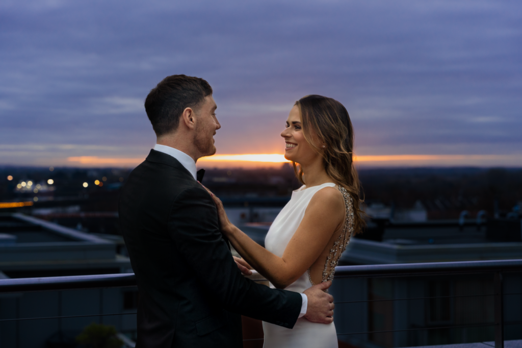 A couple dressed formally stands on a rooftop at sunset, facing each other and smiling, with city buildings visible in the background.