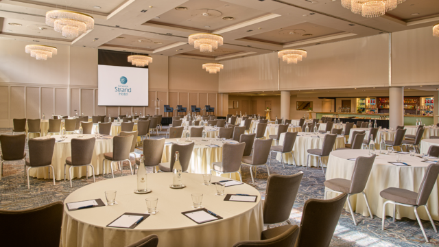 A spacious hotel conference room with round tables set for an event, notepads and water bottles at each seat, a stage, and a projector screen displaying 