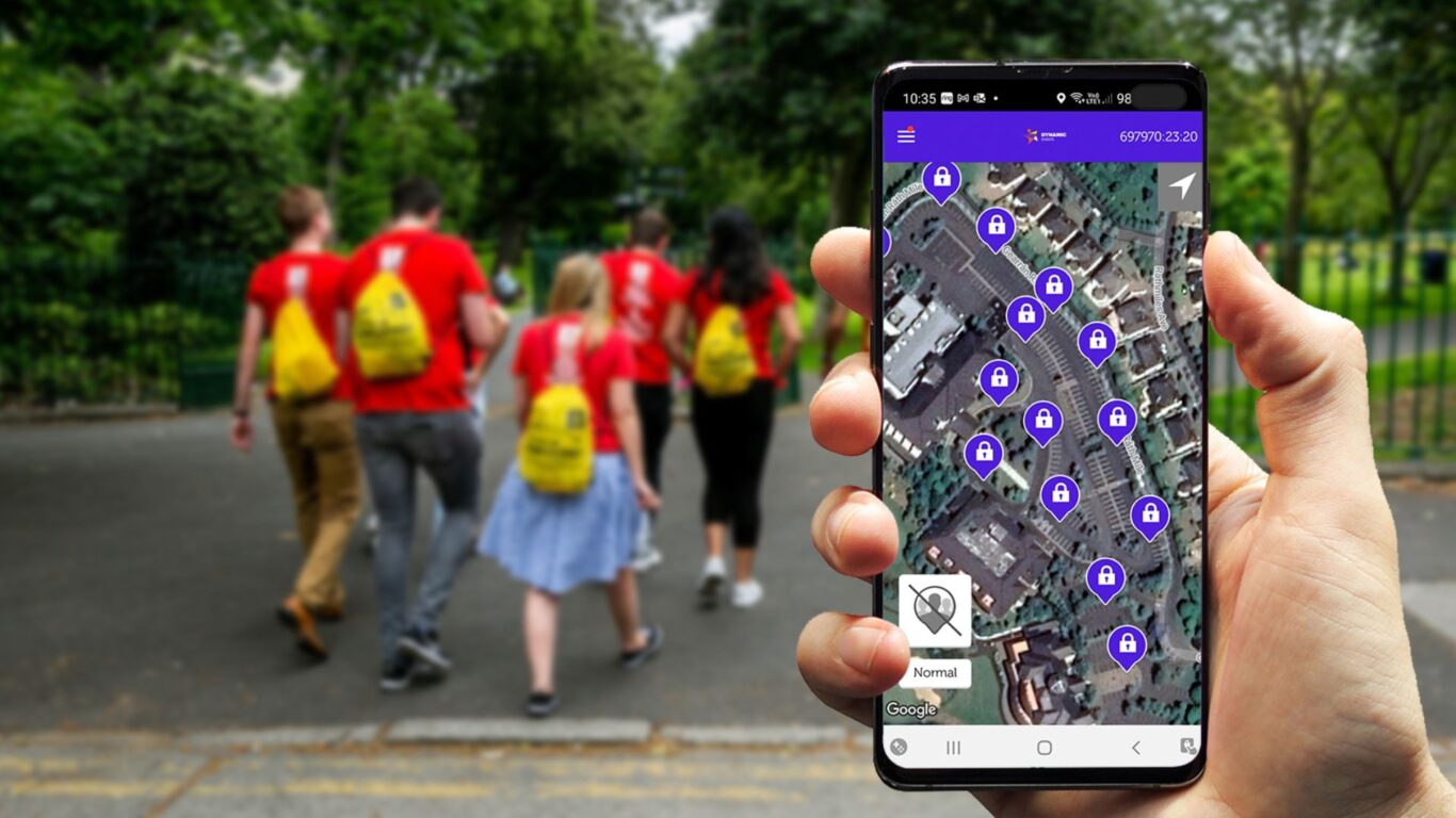 A person holds a smartphone displaying a map with lock icons, while a group of people with yellow backpacks walks ahead in a park.