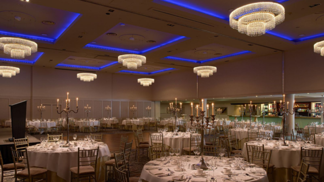 A banquet hall set for an event with round tables, white tablecloths, elegant place settings, candelabras, and chandeliers with blue ceiling lighting.