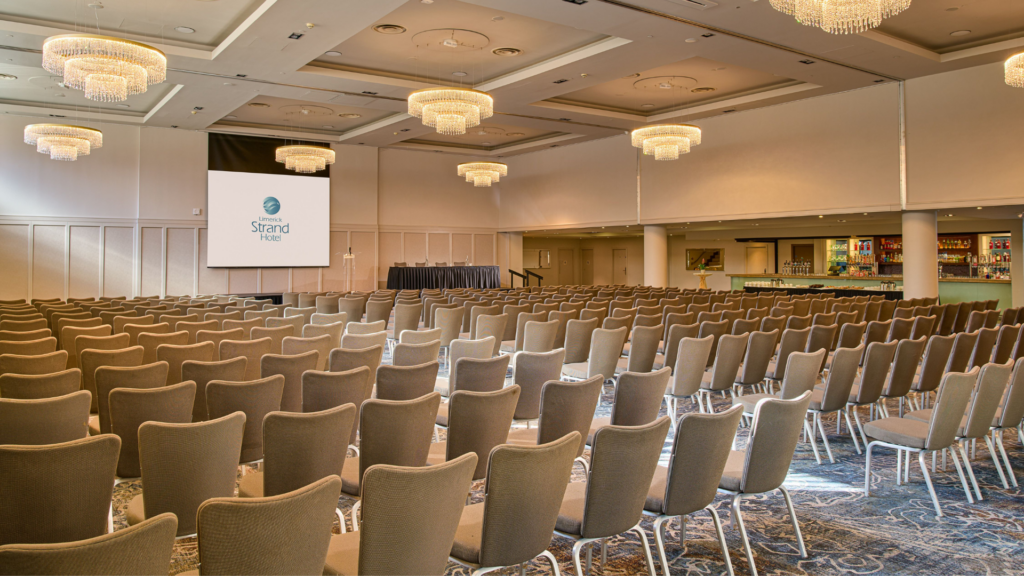 Large conference room with rows of empty chairs facing a stage and projector screen, chandeliers overhead, and a bar area visible on the right side.