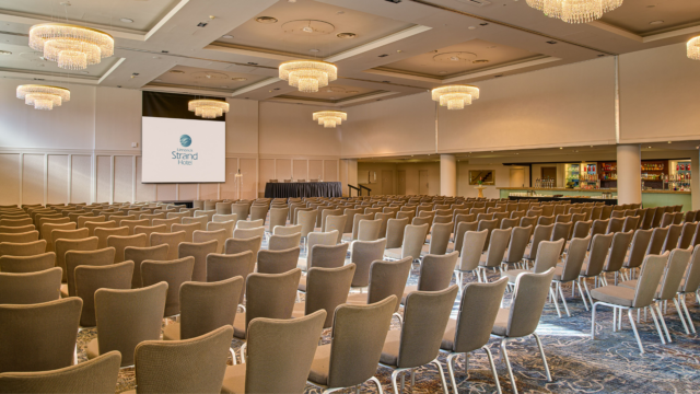 Large conference room with rows of empty chairs facing a stage and projector screen, chandeliers overhead, and a bar area visible on the right side.