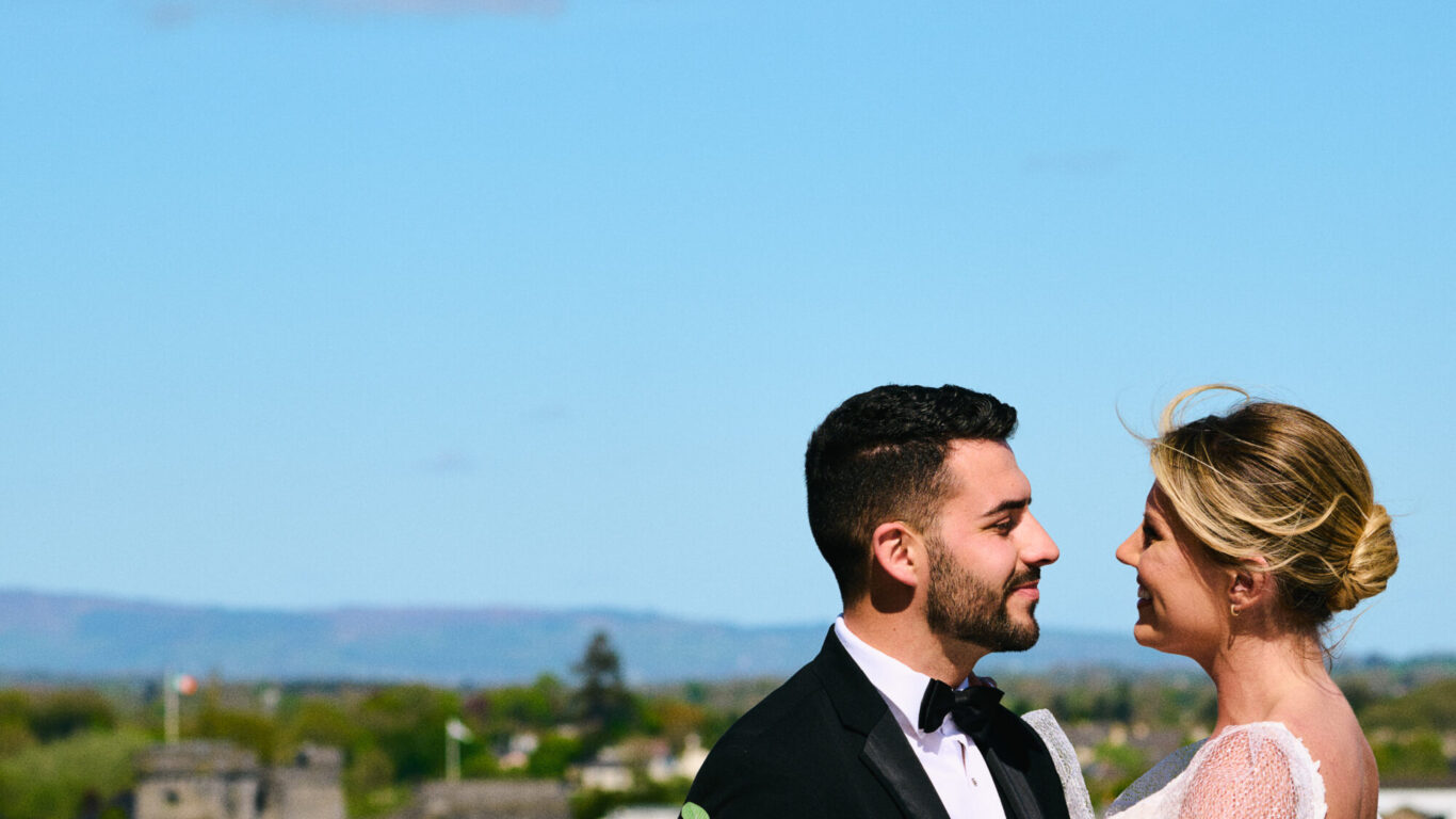 A bride and groom stand together on a balcony, holding a bouquet of white flowers, with a cityscape and blue sky in the background.