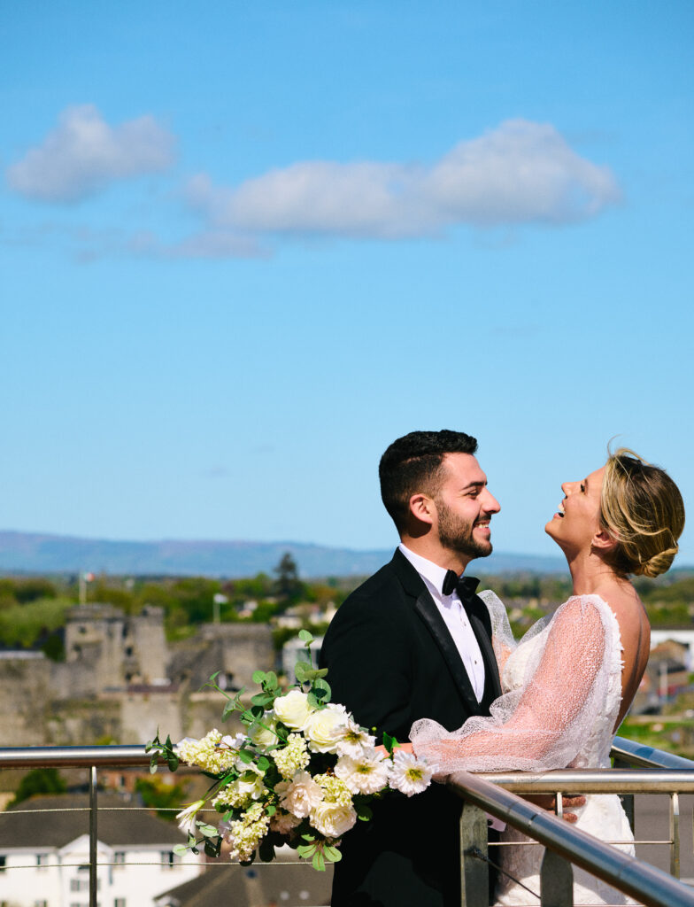 A bride and groom stand on a balcony, smiling at each other, with a bouquet of white flowers and a cityscape in the background under a clear blue sky.