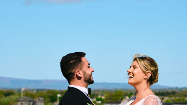 A bride and groom stand on a balcony, facing each other and smiling, with the bride holding a bouquet. There is a blue sky and distant buildings in the background.