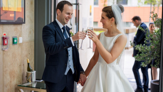A bride and groom hold hands and toast with champagne glasses indoors, both smiling, with a bottle of champagne on a table nearby.