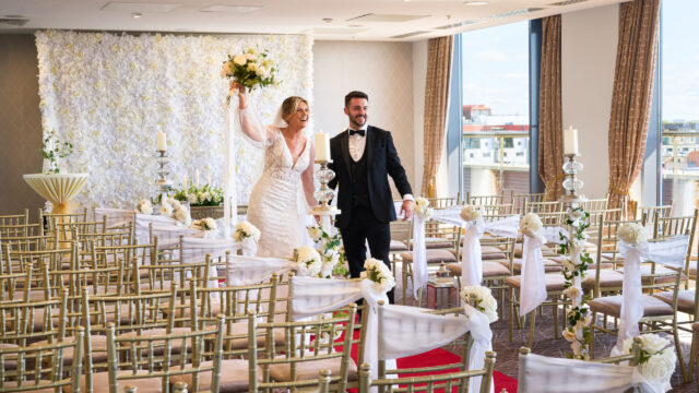 A bride and groom walk down the aisle in an indoor wedding venue decorated with gold chairs, white flowers, and large windows.